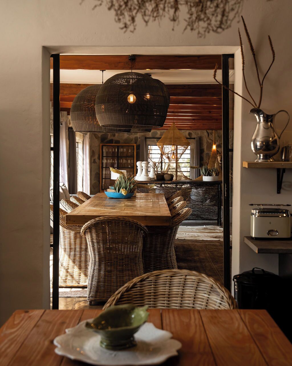 A dining area with a wooden table and wicker chairs, a large hanging light fixture, and a view into a kitchen with stone walls and wooden beams.