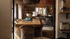 Een eetruimte met een houten tafel en rieten stoelen, een grote hanglamp en uitzicht op een keuken met stenen muren en houten balken.