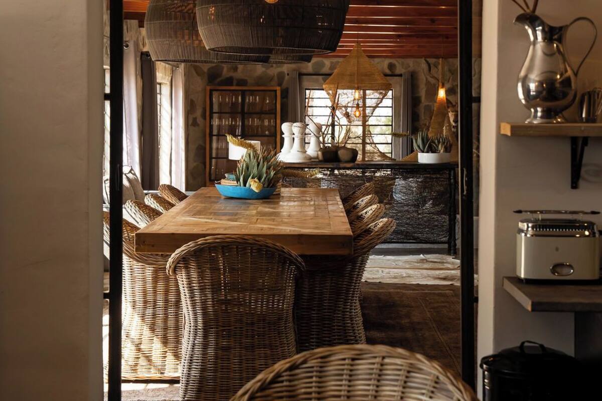 Un comedor con mesa de madera y sillas de mimbre, una lámpara colgante grande y vista hacia una cocina con paredes de piedra y vigas de madera.