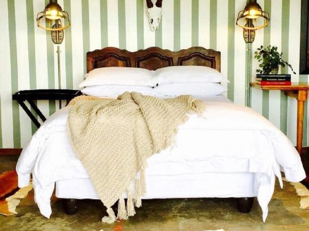 Ein Bett mit Holz-Kopfteil, weißer Bettwäsche und einer beigen Decke. Zwei Wandlampen und ein kleiner Tisch mit einer Pflanze sind vorhanden.