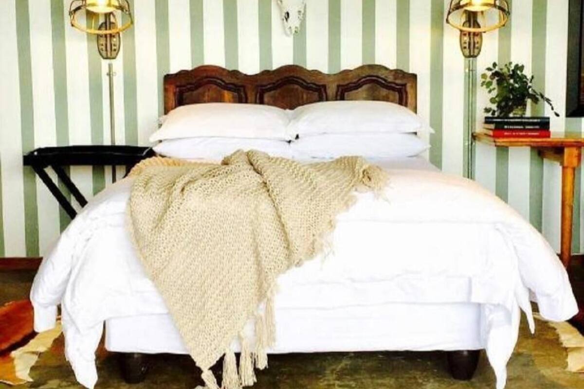 Una cama con cabecera de madera, ropa de cama blanca y una manta color beige. Hay dos lámparas fijadas a la pared y una mesita con una planta.