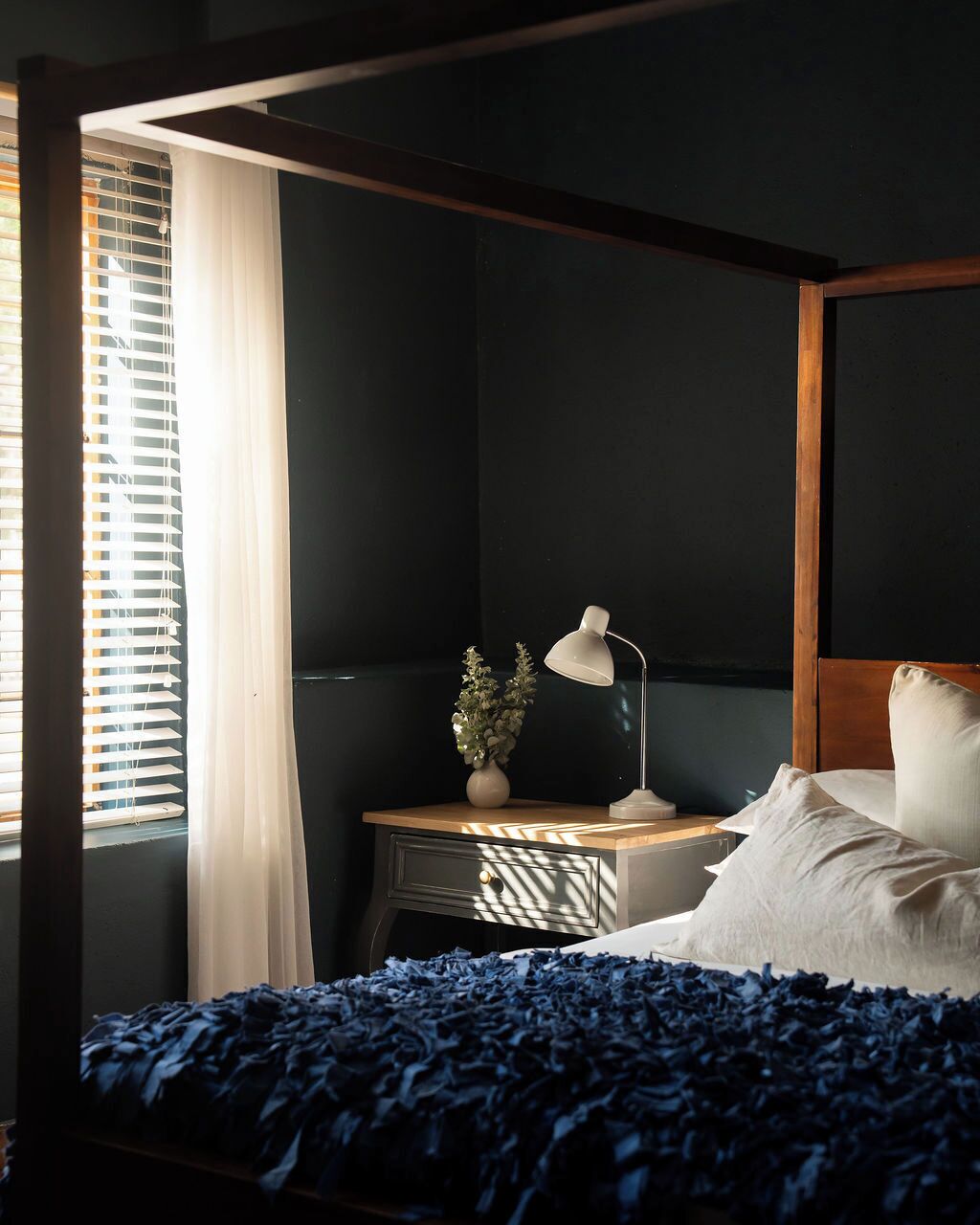 A bedroom with a bed, a nightstand, a lamp, a plant, and a window with blinds.