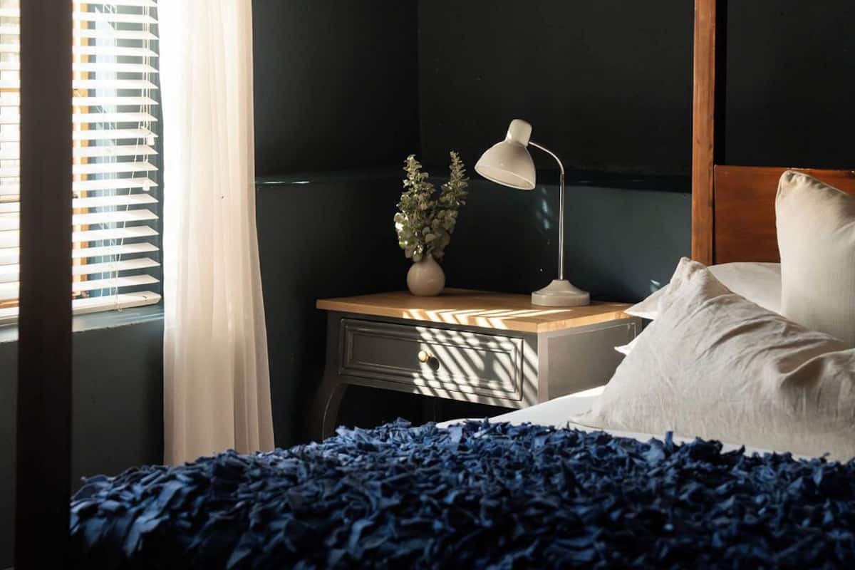 Un dormitorio con cama, mesita de noche, lámpara, planta y ventana con persianas.