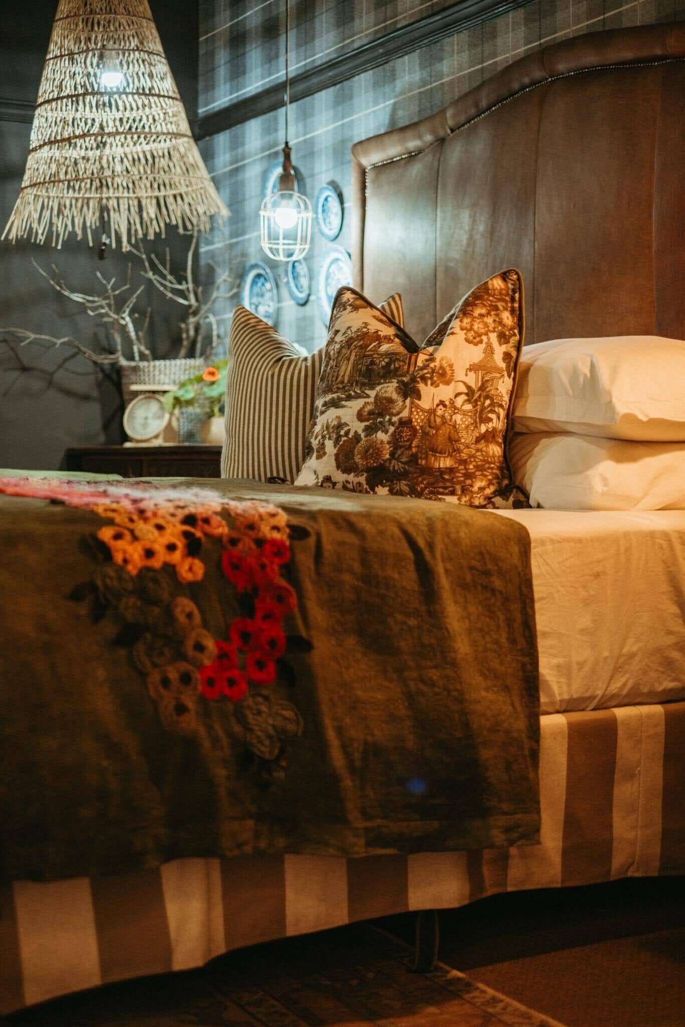A bed with a striped bedspread, decorative pillows, a hanging woven light fixture, and wall-mounted lanterns.