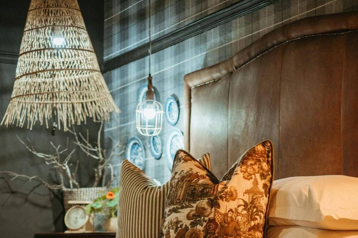 Una cama con colcha a rayas, almohadas decorativas, una lámpara colgante tejida y faroles fijados a la pared.