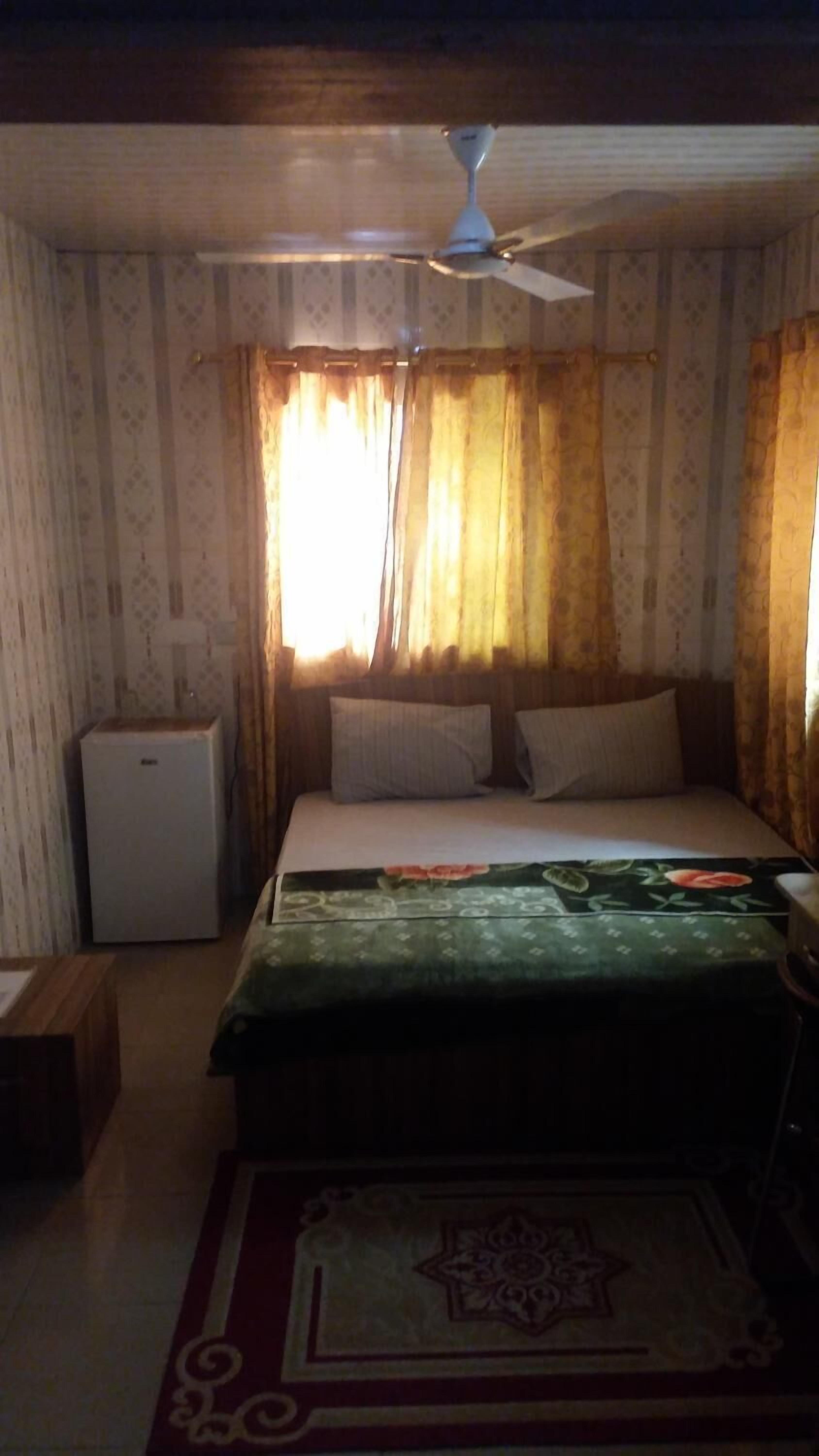 Un dormitorio con cama, ventilador de techo, un refrigerador pequeño y cortinas.