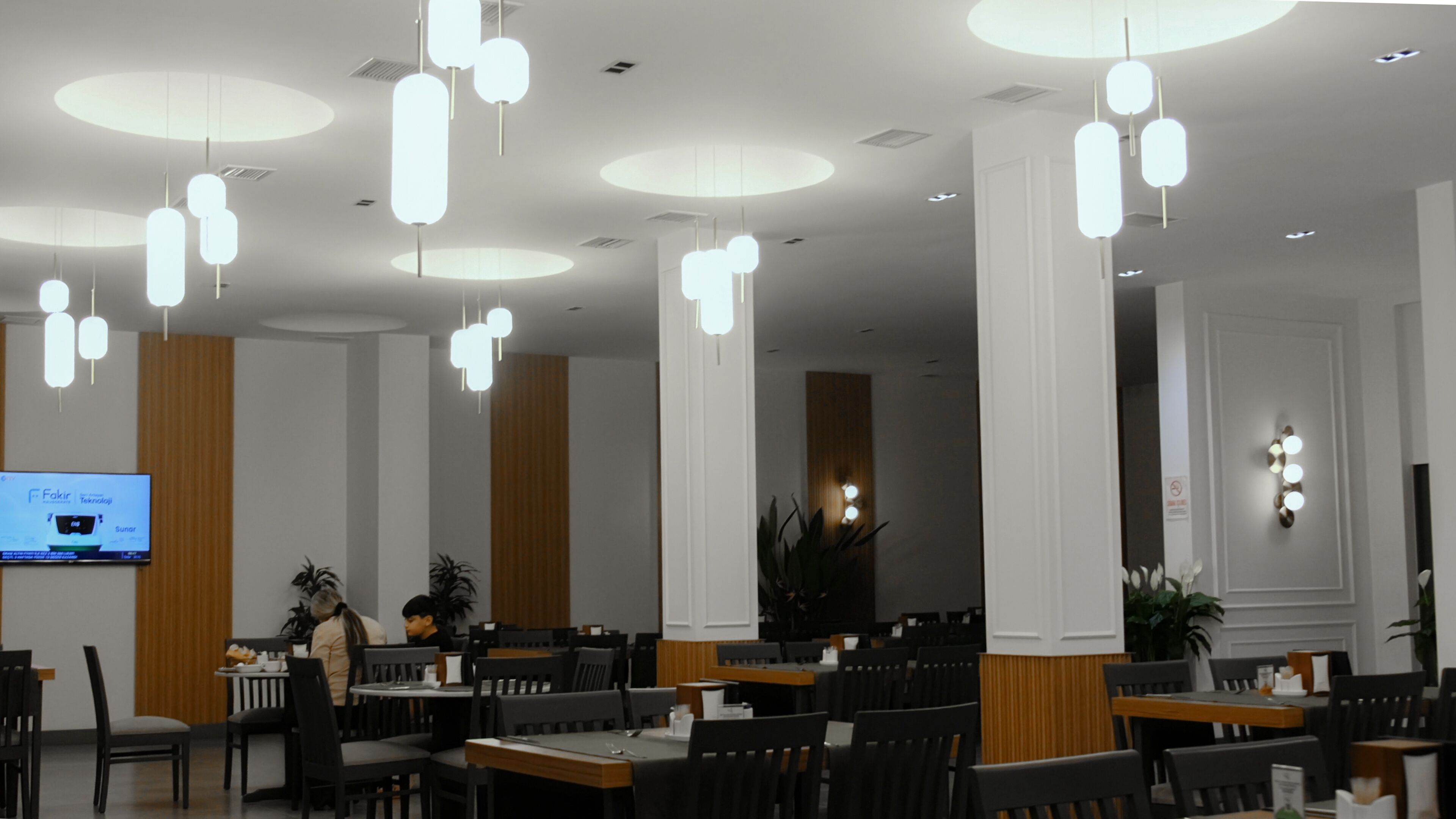 Un restaurant doté de nombreuses tables et chaises, d’un éclairage moderne avec des suspensions et d’un écran de télévision diffusant du contenu.