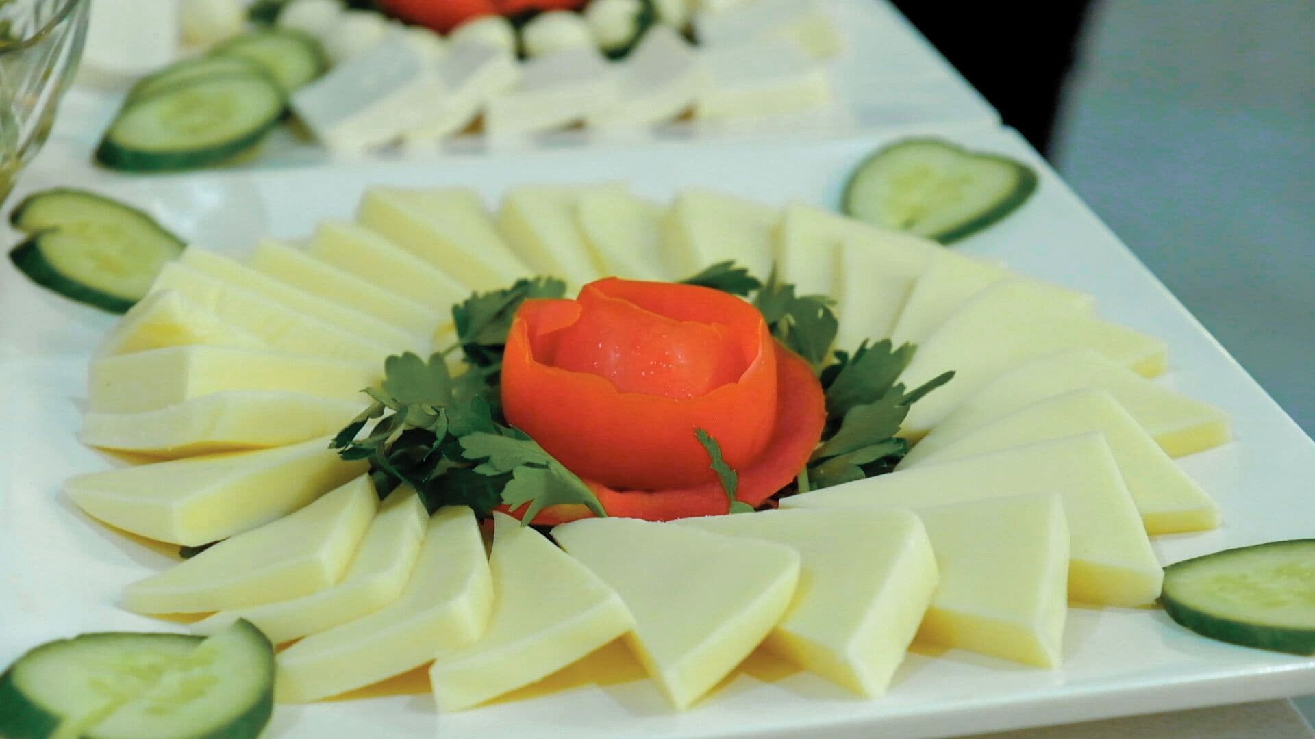 A white plate with cheese slices arranged in a circular pattern around a central red rose, with cucumber slices on the side.