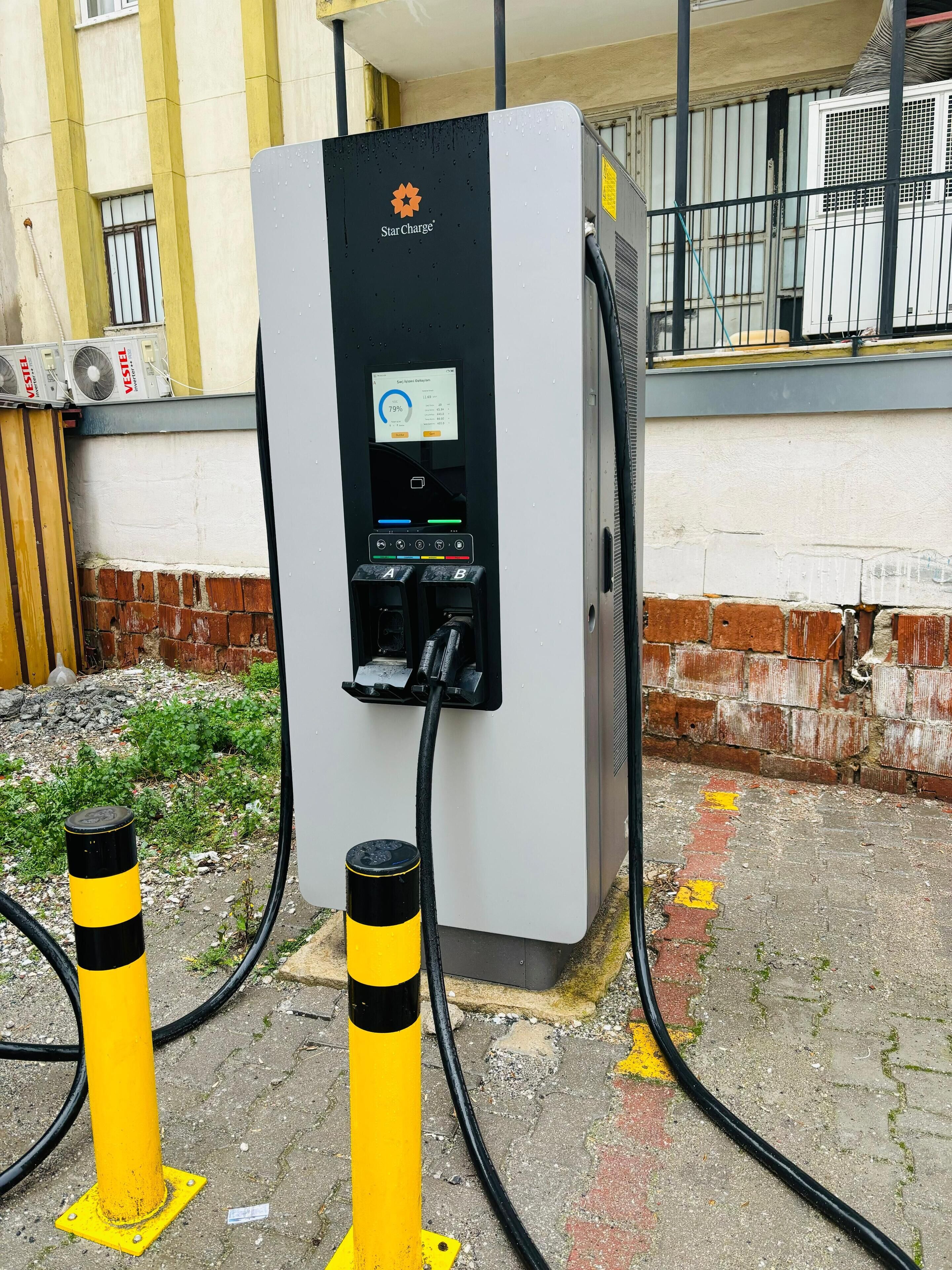 Una estación de carga para vehículos eléctricos Star Charge con pantalla digital, conectada a un edificio con unidades de aire acondicionado y una pared de ladrillo.