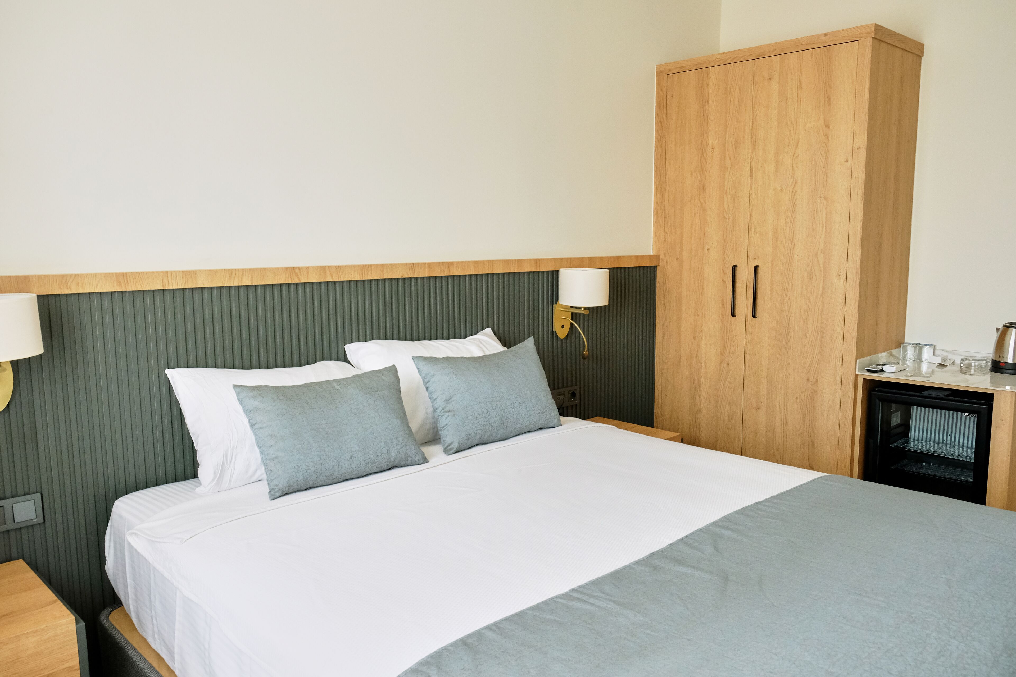 A neatly made bed with white and grey linens, a wooden wardrobe, and a small bedside table with a lamp.