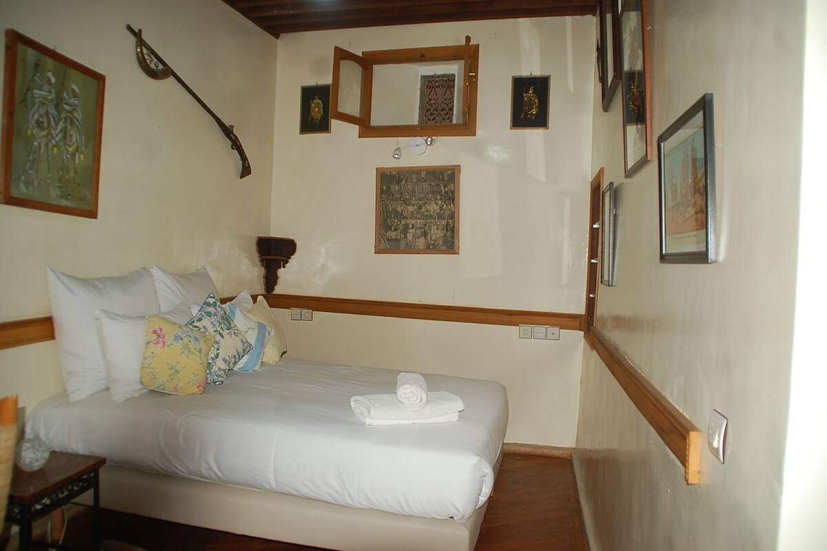 A hotel room with a bed, pillows, a small table, and a wall with framed pictures and a mirror.