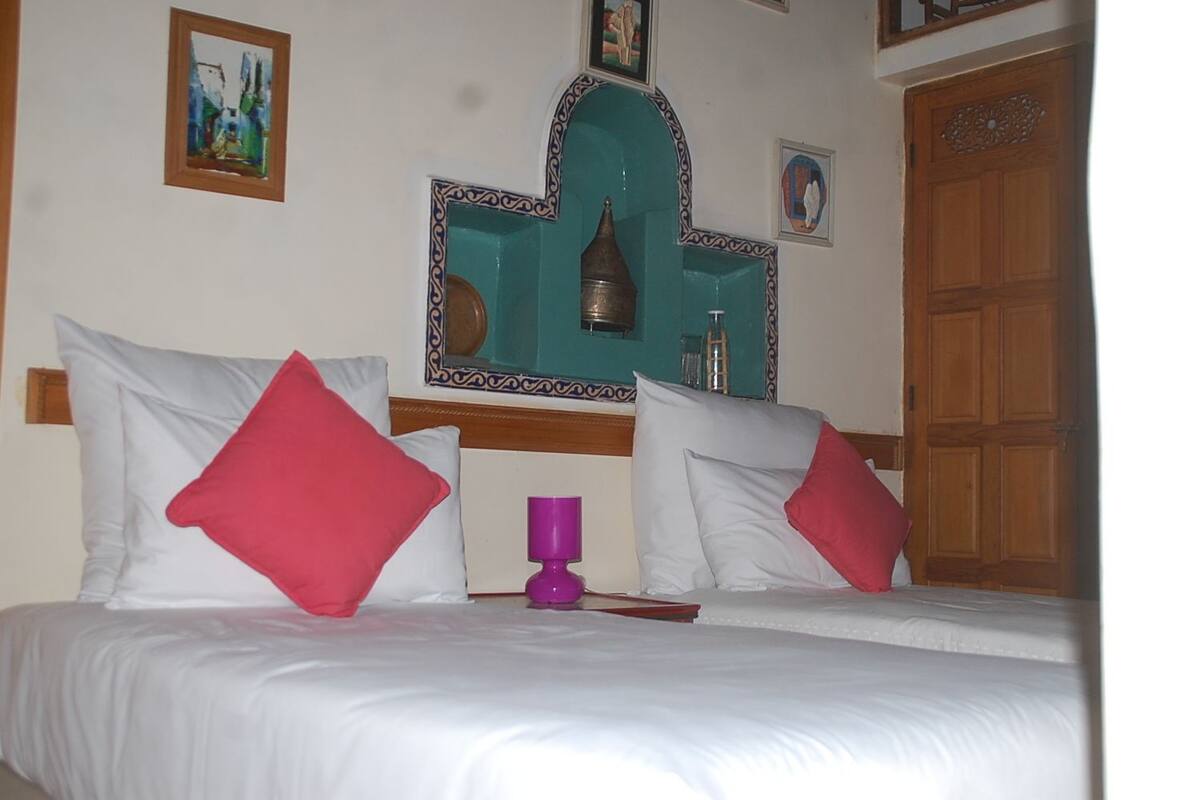 A hotel room with a bed, pillows, a bedside table, a mirror, and a wooden door.