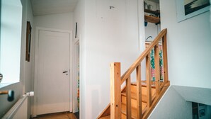 A wooden staircase with a handrail leading to a landing. A white door is visible to the left, and a window with blinds is on the left wall. - Bessastadir Guesthouse (Hvammstangi)