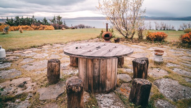 A rustic wooden table with a circular top, surrounded by stone slabs and wooden posts, set in a natural landscape with a body of water and mountains in the background. - Bessastadir Guesthouse (Hvammstangi)