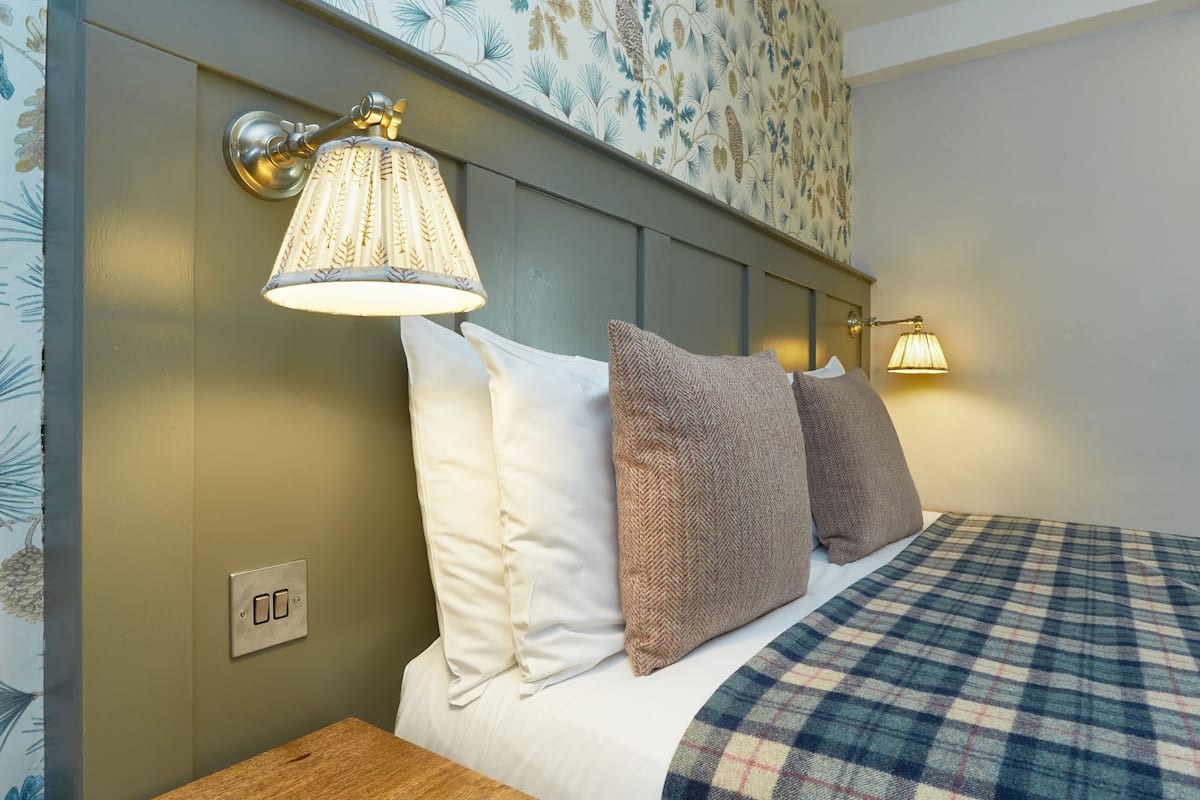 Una cama bien hecha con ropa de cama escocesa, dos lámparas fijadas a la pared y un estampado floral en el papel tapiz sobre la cama.