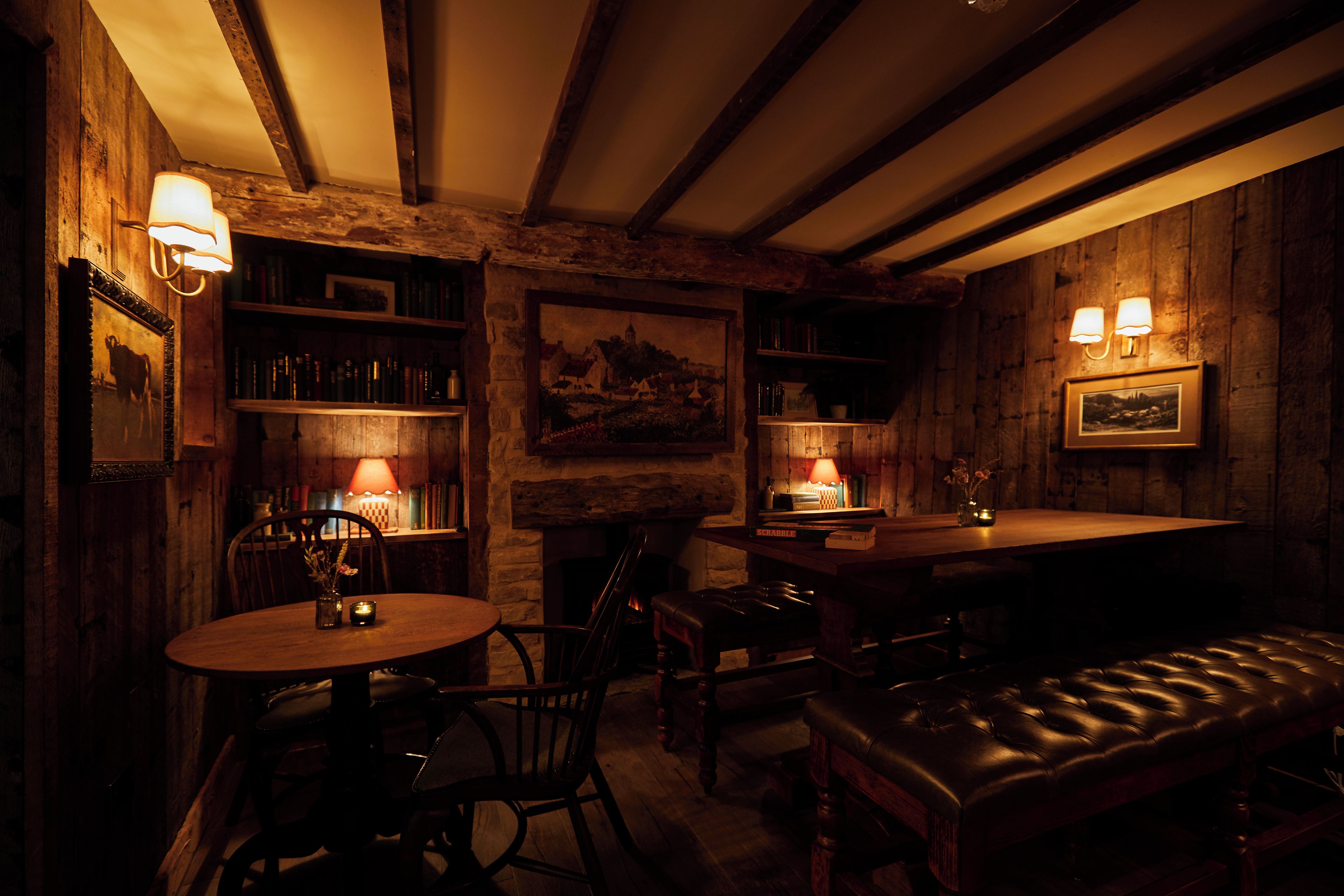 A dimly lit room with wooden walls, a fireplace, a round table, a bench, and bookshelves with lamps.