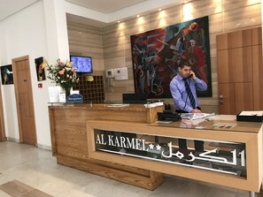 A reception desk with a wooden finish and a glass partition. There is a television mounted on the wall above the desk. - Hotel Al Karmel (Tunis)