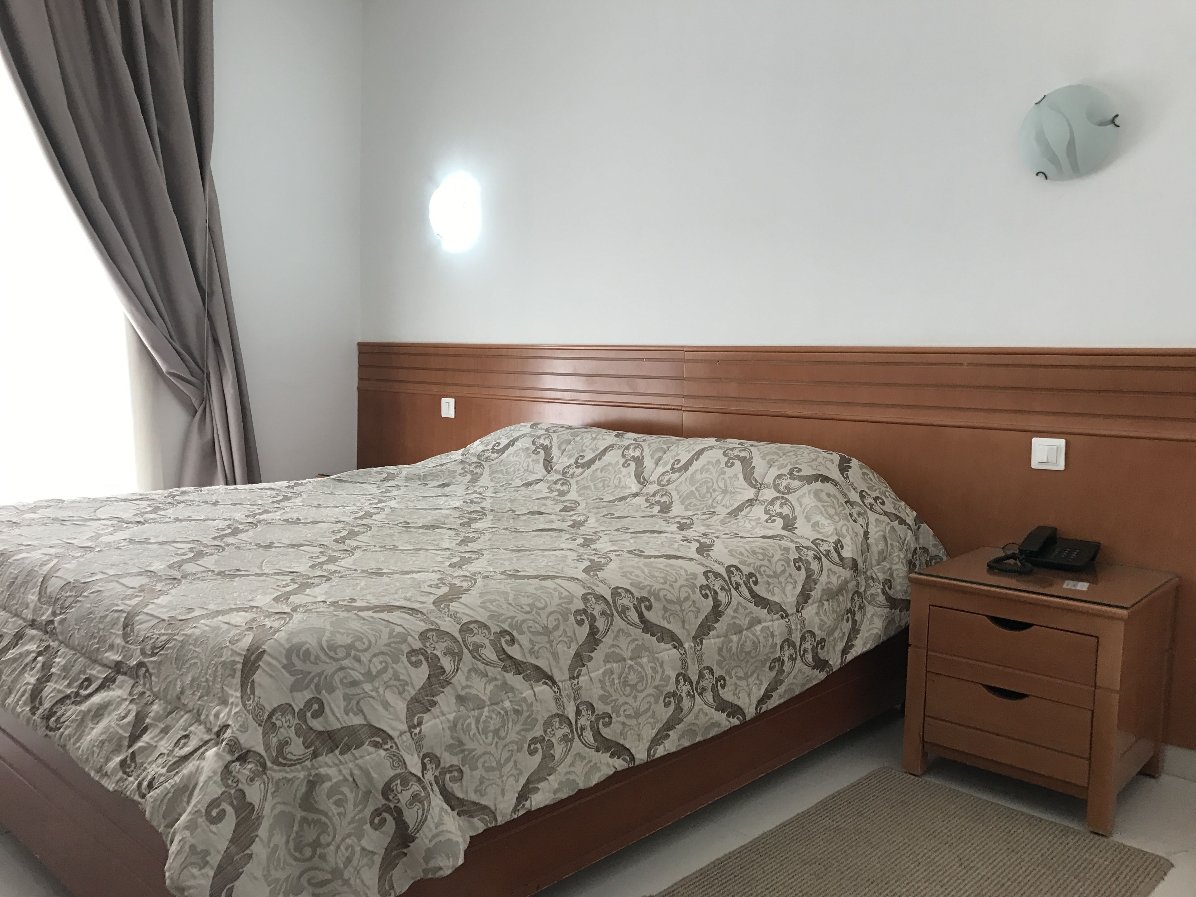Un dormitorio con cama, mesita de noche, reloj de pared y toma de corriente.