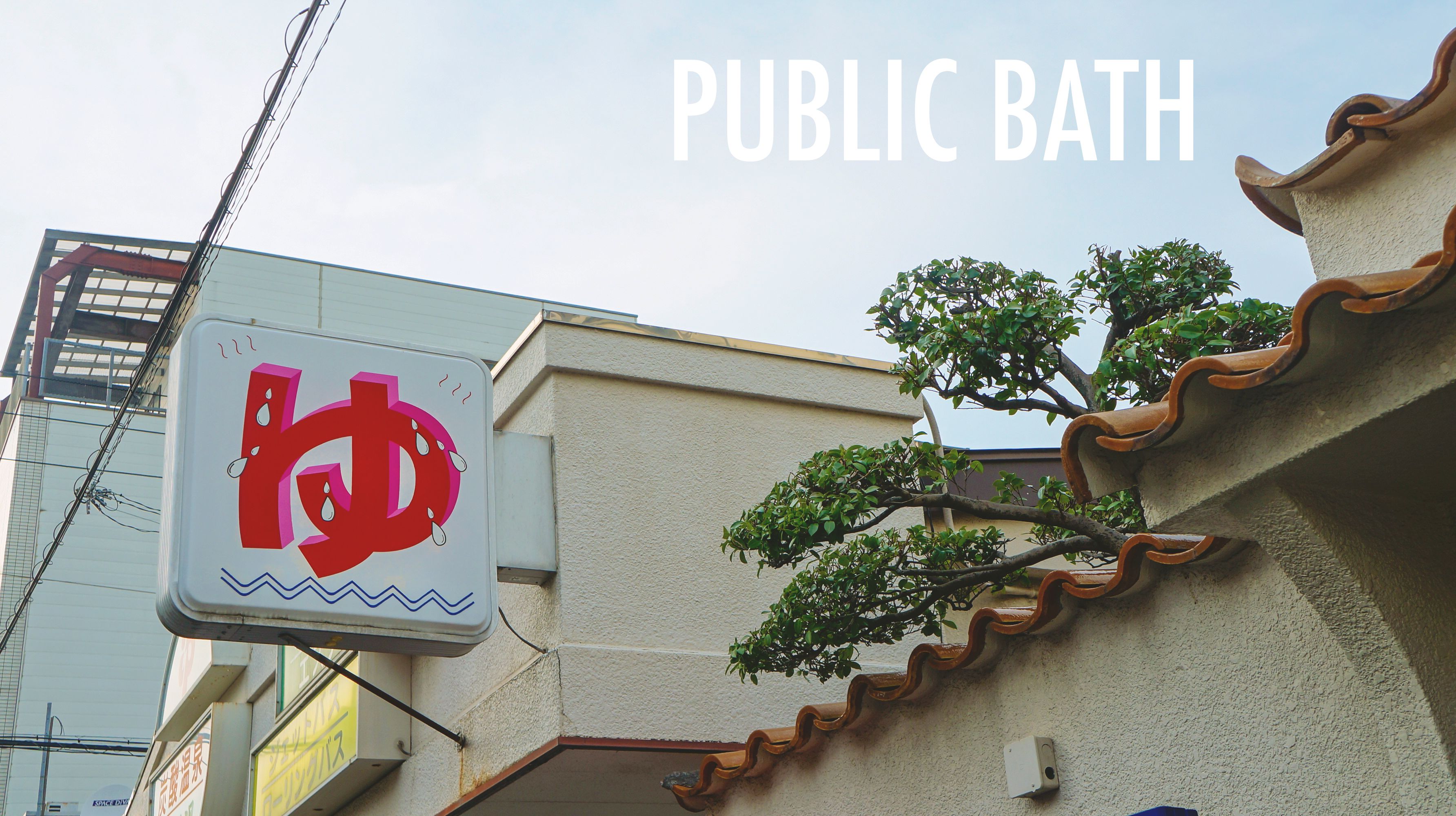 A public bath sign with a stylized letter 'D' and droplet design, mounted on a building with a tiled roof and a tree in the foreground.