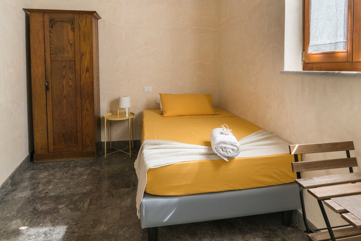 Quarto pequeno com uma cama de solteiro, guarda-roupa de madeira, mesinha de cabeceira com abajur e janela com moldura de madeira.