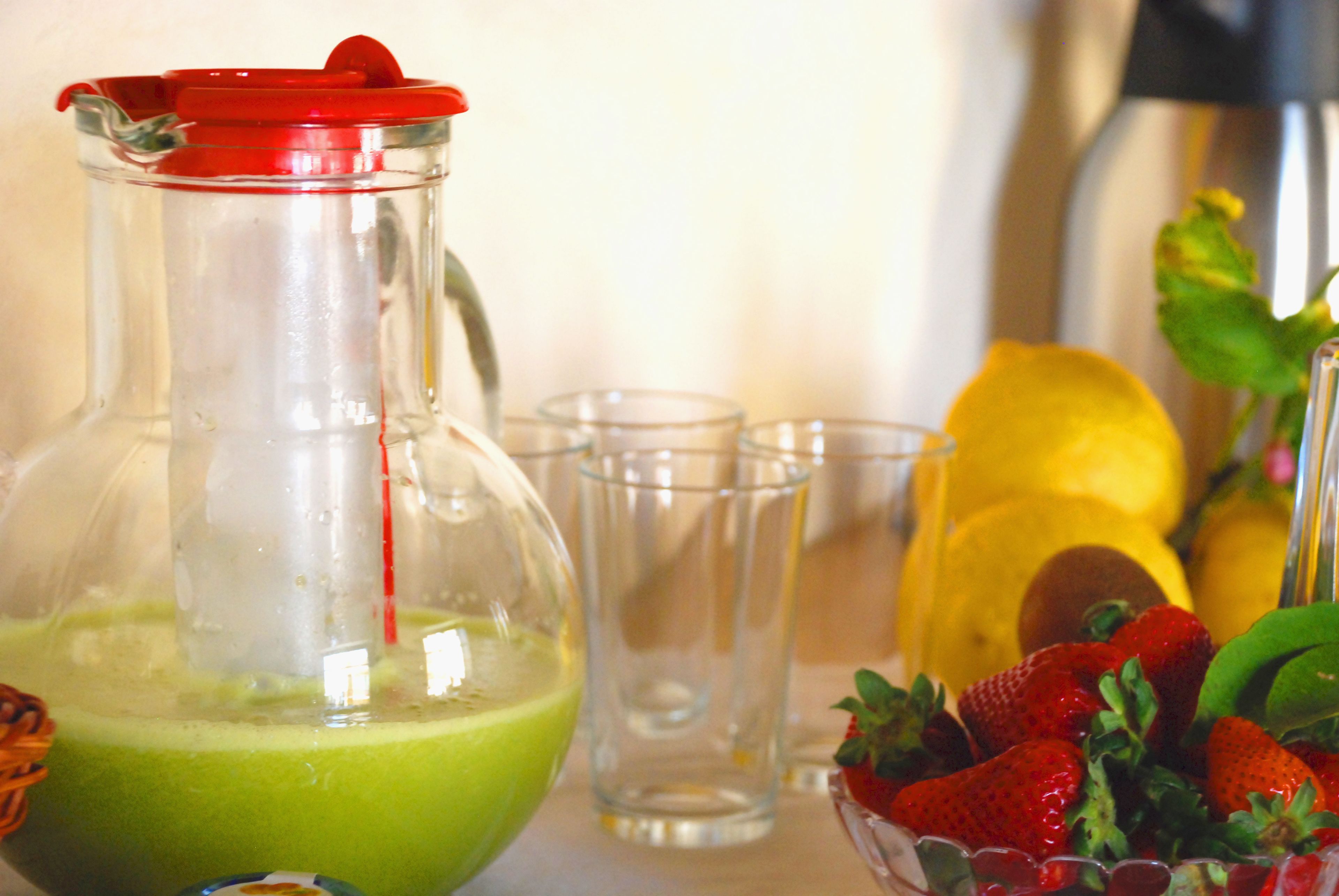 Um jarro de smoothie verde, uma tigela de morangos e uma fruta amarela, possivelmente um limão, sobre uma mesa.