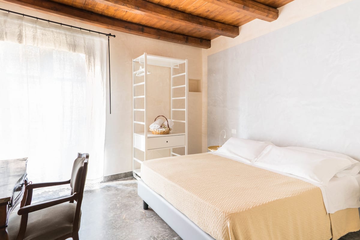 Quarto com cama, uma cadeira de madeira, guarda-roupa branco e janela com cortinas.
