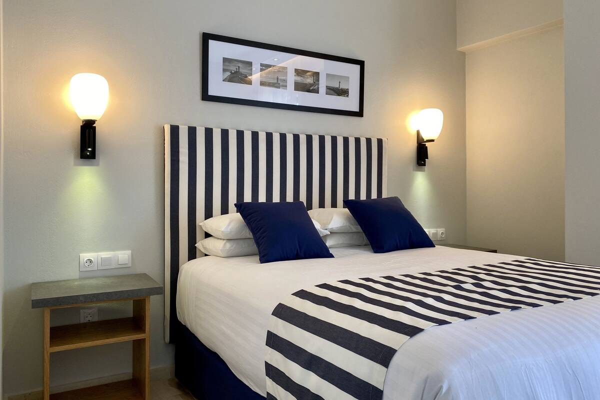 A hotel room with a striped bedspread, a headboard with vertical stripes, two wall-mounted lights, a bedside table, and a framed picture above the bed.