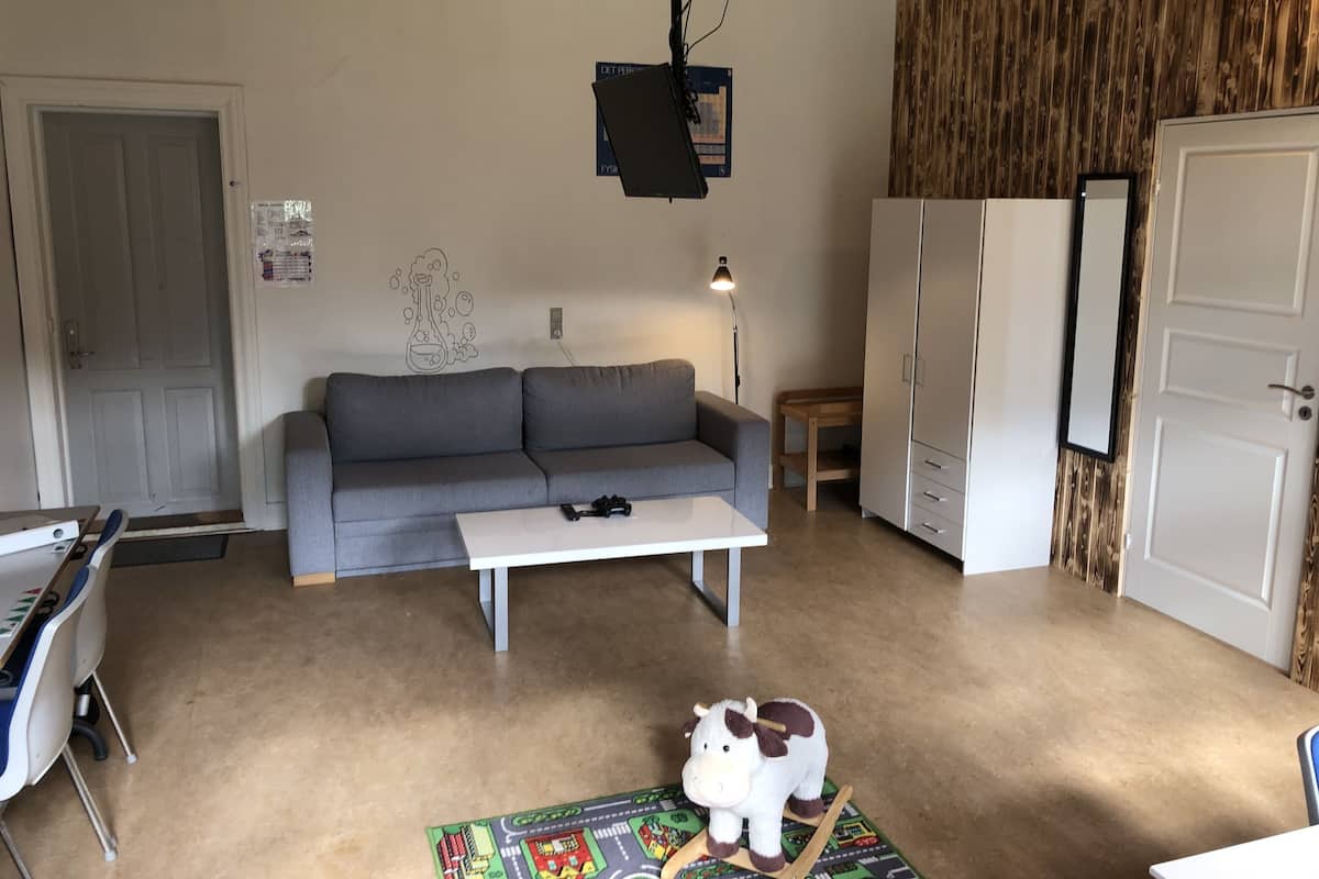 A living room with a grey sofa, a white coffee table, a small wooden table, a lamp, a white wardrobe, and a colorful rug.