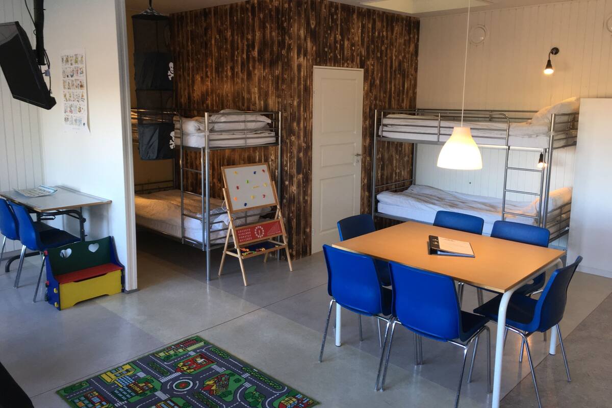 A dormitory room with bunk beds, a study table, blue chairs, a colorful rug, and a wooden accent wall.