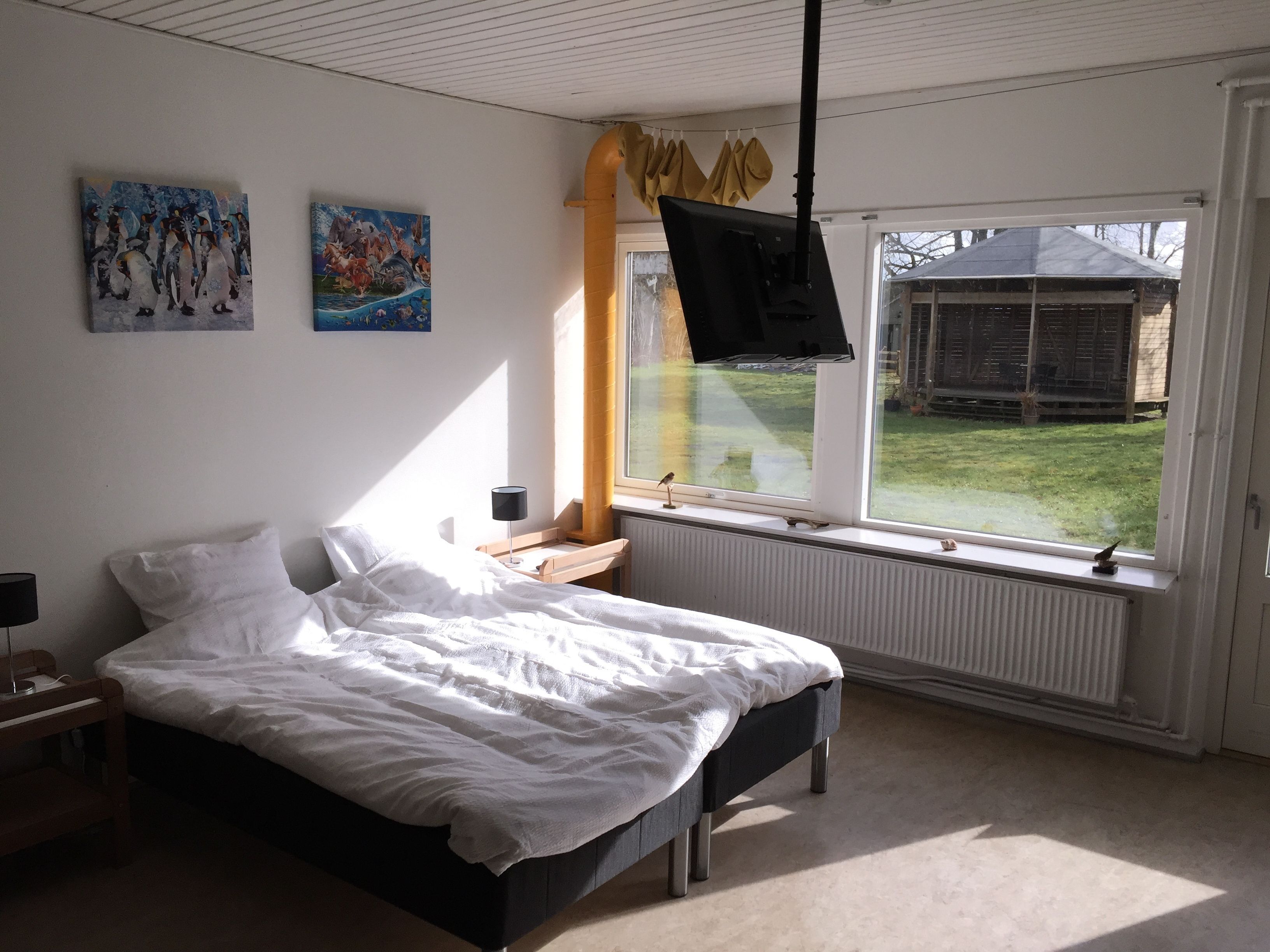 Et soveværelse med en seng, et TV ophængt på væggen, et vindue med gardiner og billedkunst på væggen.