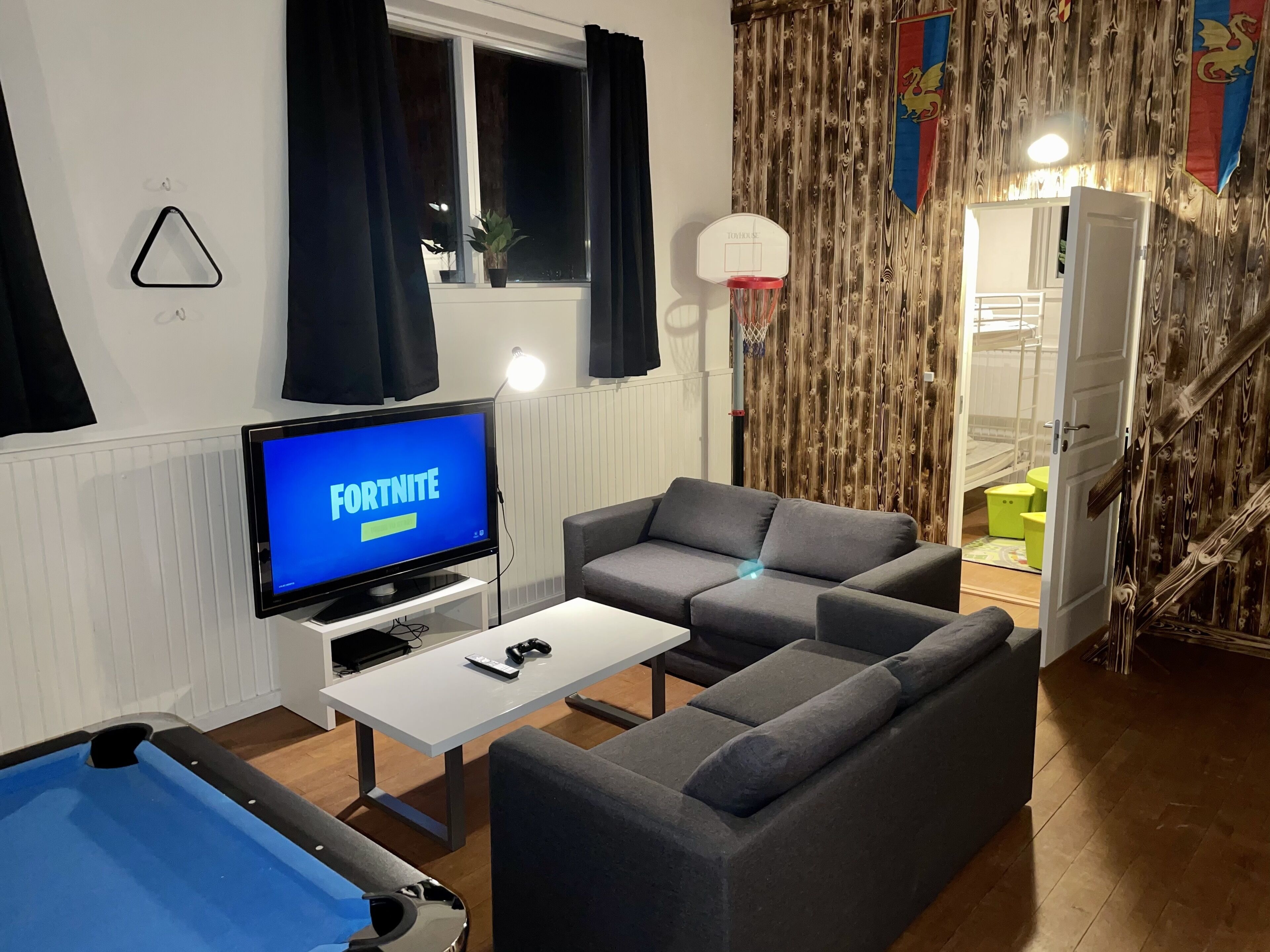 En stue med et fjernsyn, der viser 'FORTNITE', en grå sofa, et billardbord og en trævæg med flag.