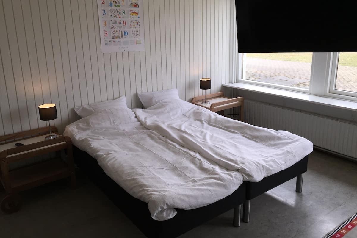 A neatly made bed with white bedding, a bedside table with a lamp, and a poster on the wall.