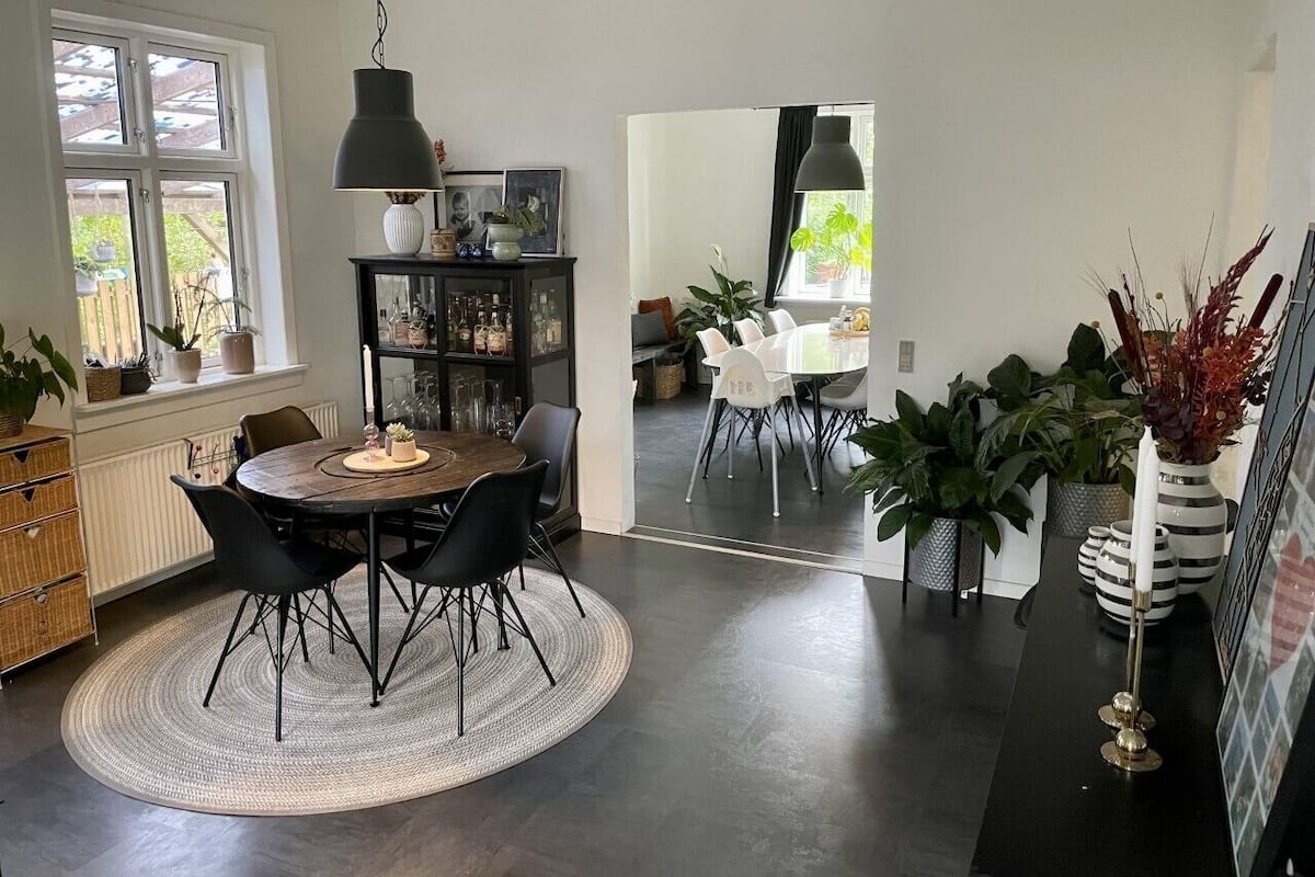 A modern dining area with a round table, black chairs, and a dark floor. There is a bookshelf with various items and a window with a view of greenery.