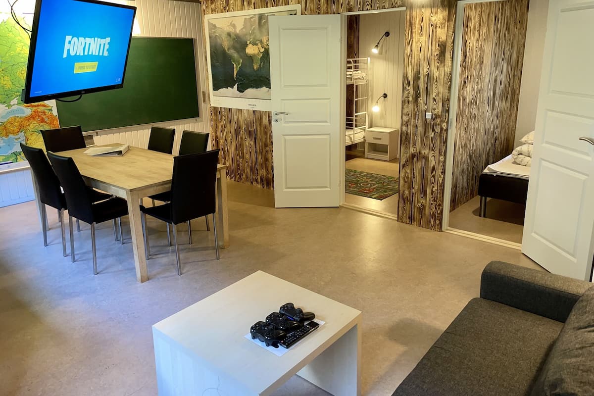 A room with a wooden wall, a desk with chairs, a television displaying 'Fortnite', and a door leading to a bedroom.