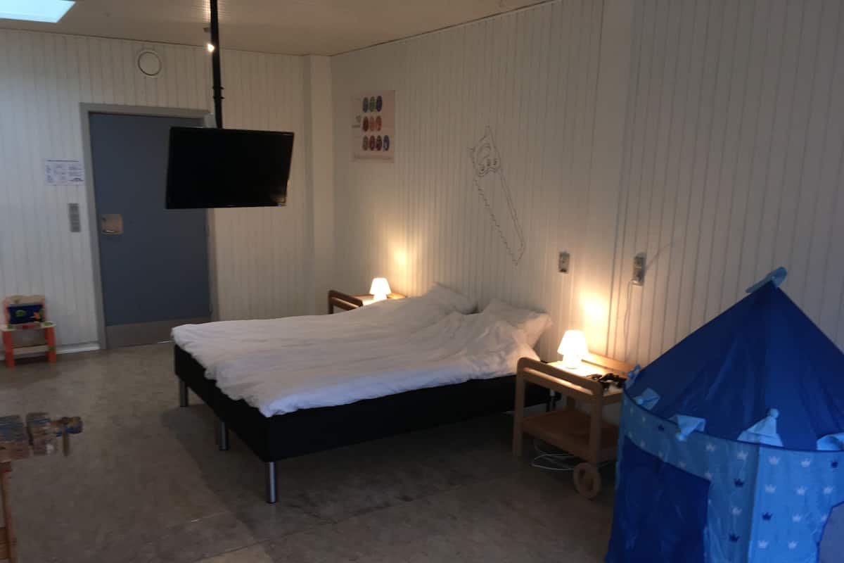 A bedroom with a bed, two bedside tables with lamps, a TV mounted on the wall, a blue tent-like structure, and a door.