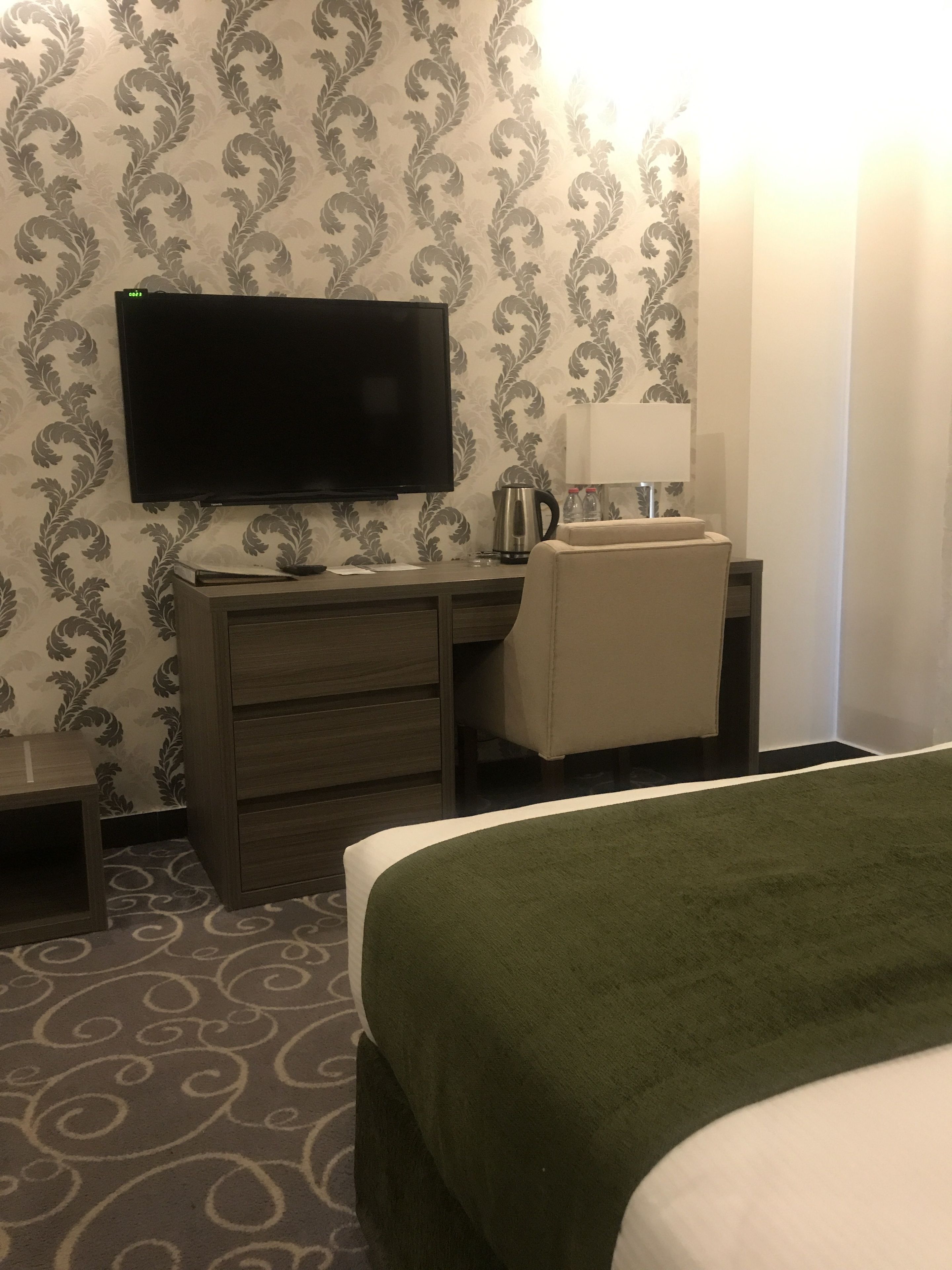 A hotel room with a bed, a desk with a flat-screen TV, a coffee maker, and a lamp.