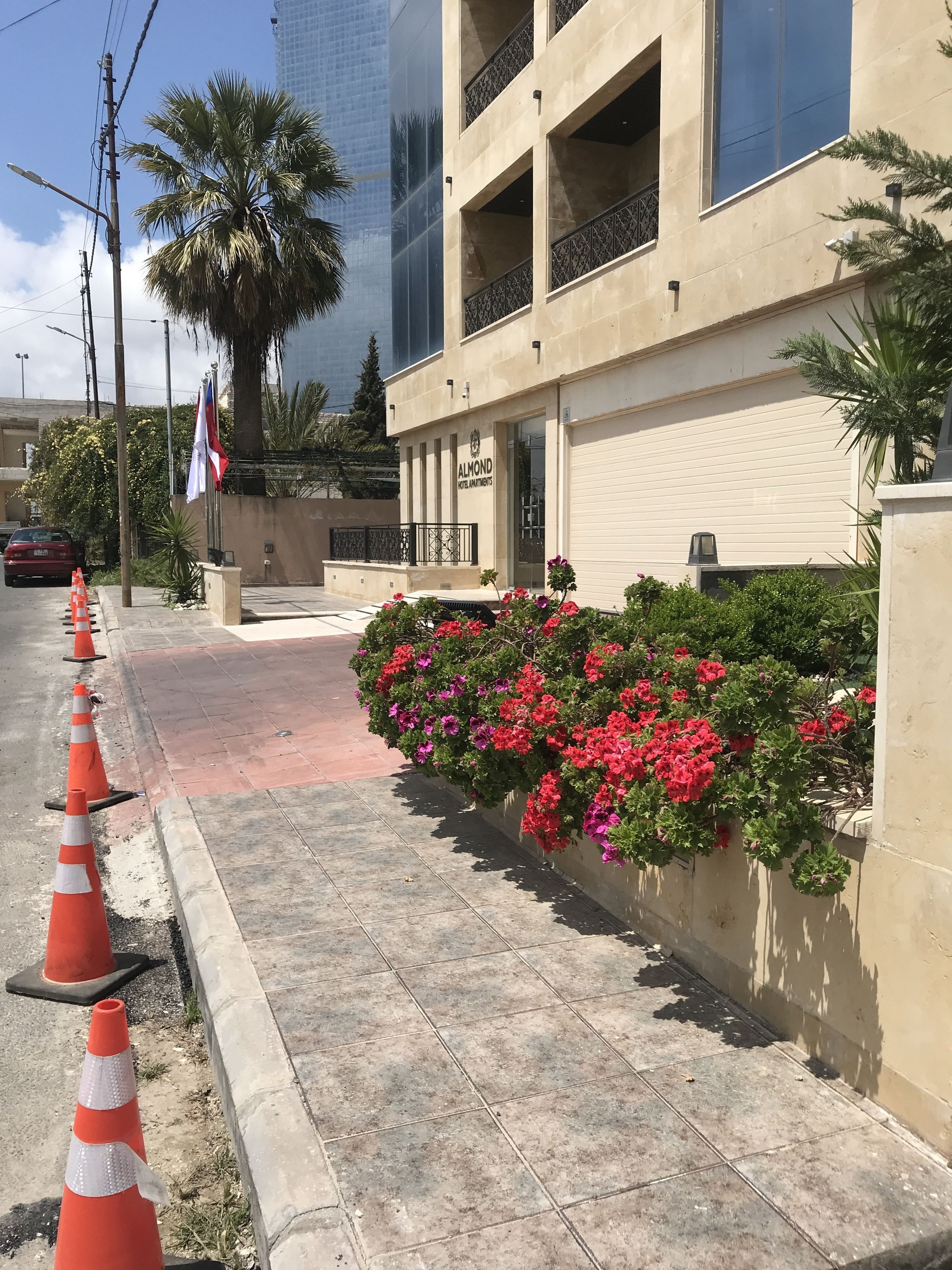 L’entrée d’un hôtel avec une façade beige, un palmier et un parterre fleuri avec des fleurs rouges.