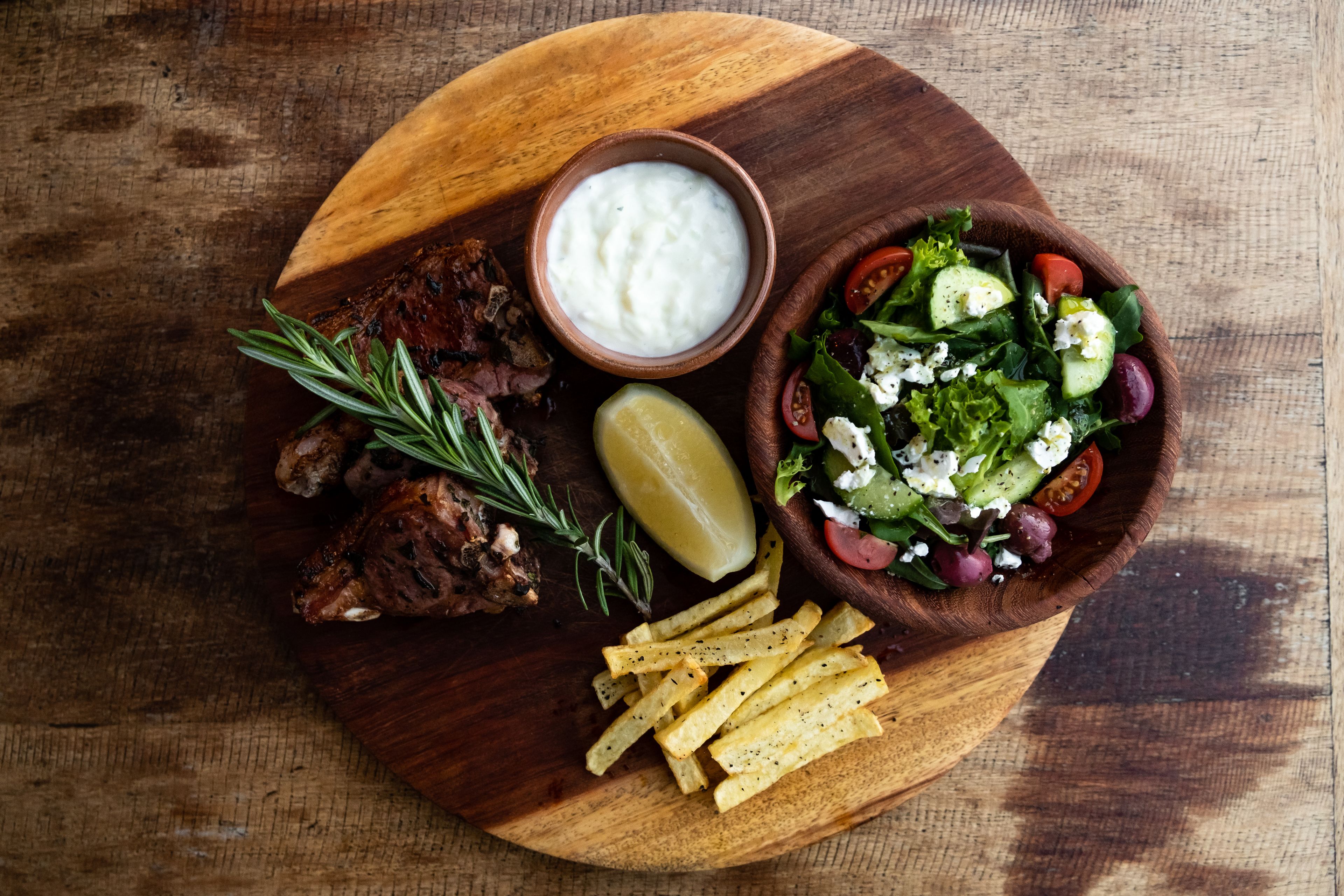 Ein Holzbruchbrett mit gegrilltem Fleisch, einer Salatschüssel mit Feta-Käse, einem Zitronenzwischenstück und Pommes Frites.
