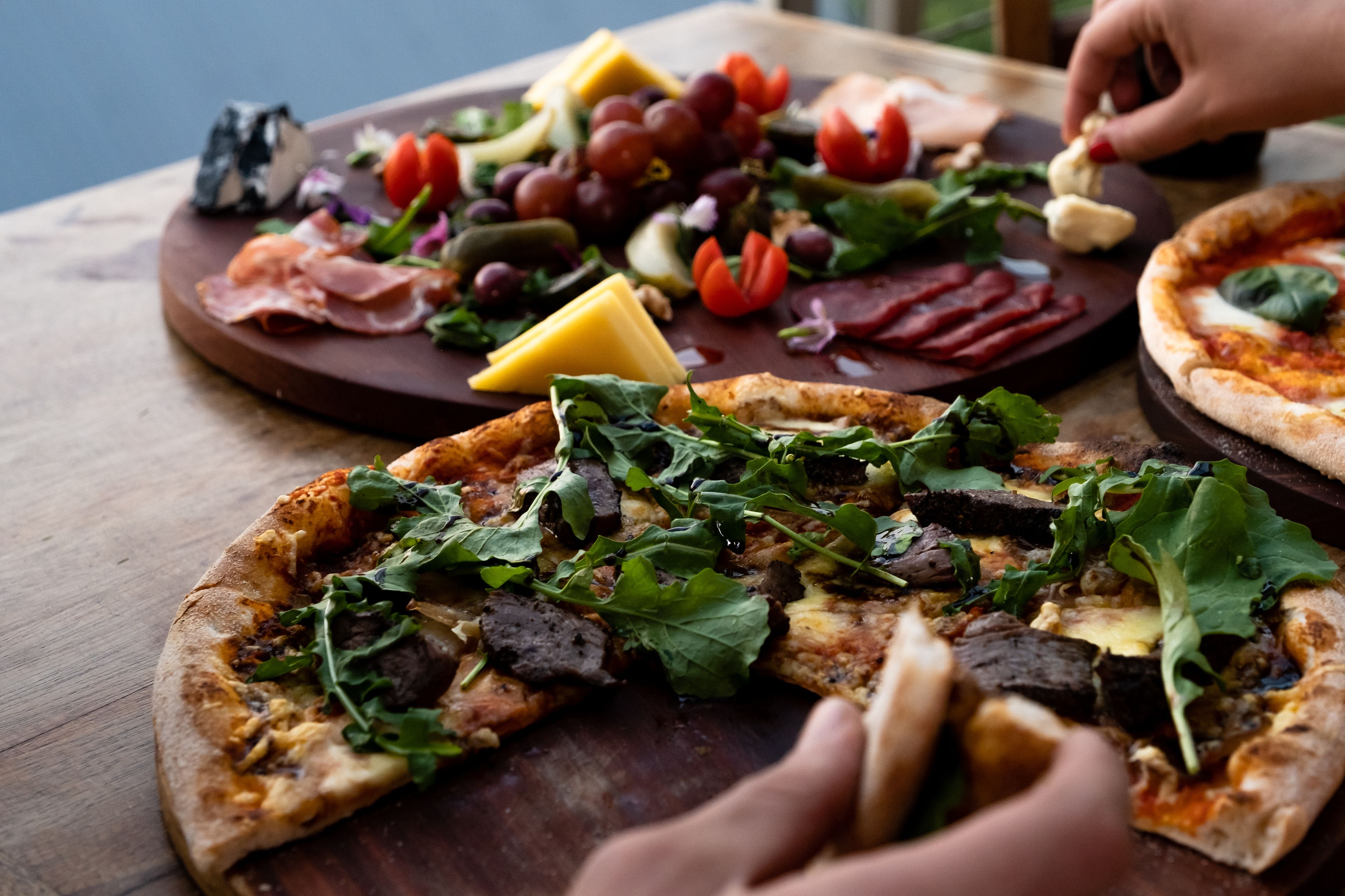 A wooden table with three pizzas topped with various ingredients and a selection of cheeses, grapes, and cured meats.