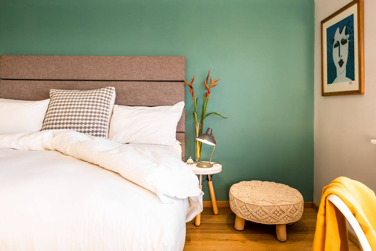 A neatly made bed with white linens and patterned pillows, a bedside lamp, a small round ottoman, and a framed artwork on the wall.