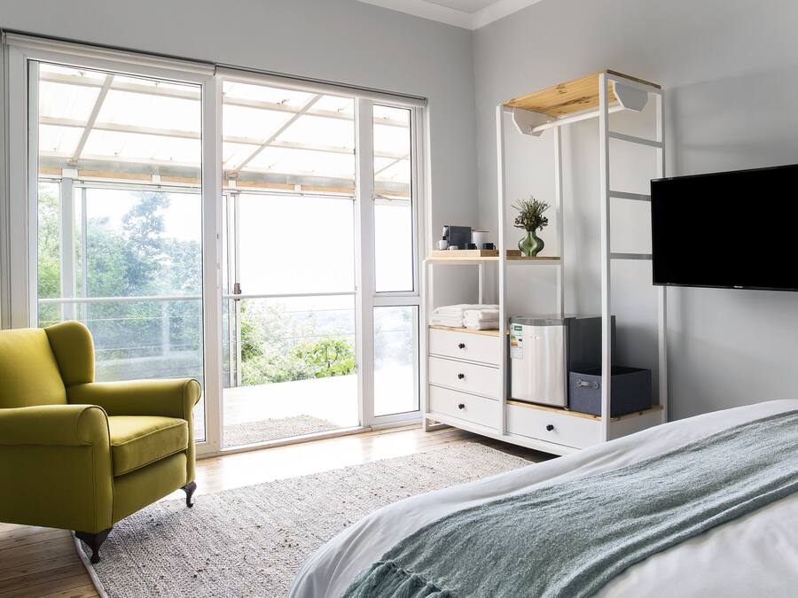 A bedroom with a bed, a green armchair, a wardrobe, a television, and a sliding glass door leading to a balcony.