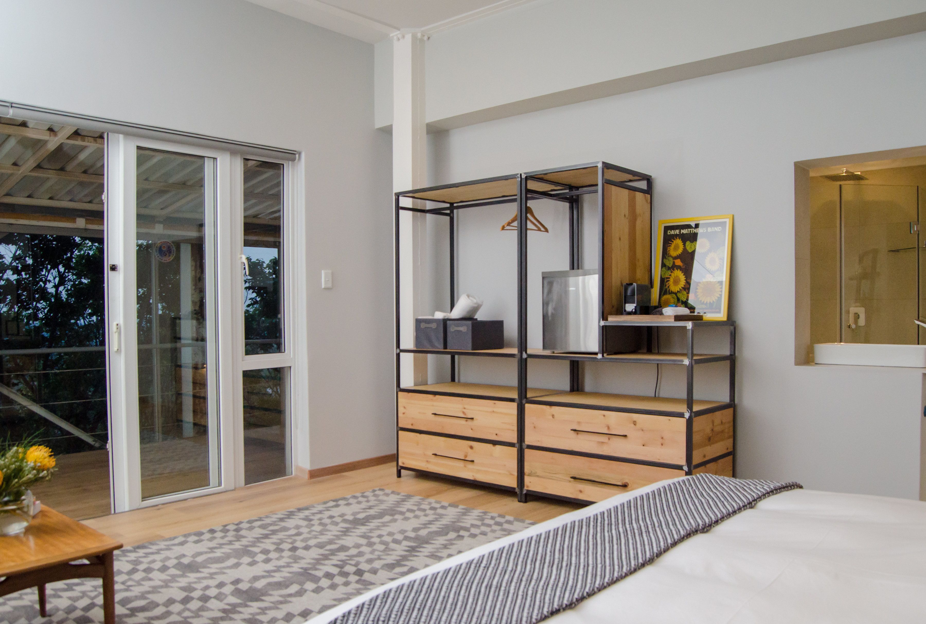A bedroom with a bed, a wooden dresser, a metal wardrobe, a bathroom mirror, and a sliding glass door leading to a balcony.