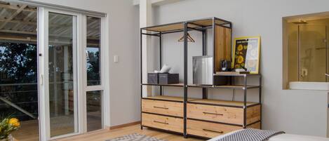 A bedroom with a bed, a wooden dresser, a metal wardrobe, a bathroom mirror, and a sliding glass door leading to a balcony.