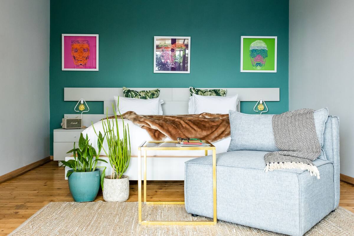 A bedroom with teal walls, a bed with white bedding, a grey sofa, and two potted plants.