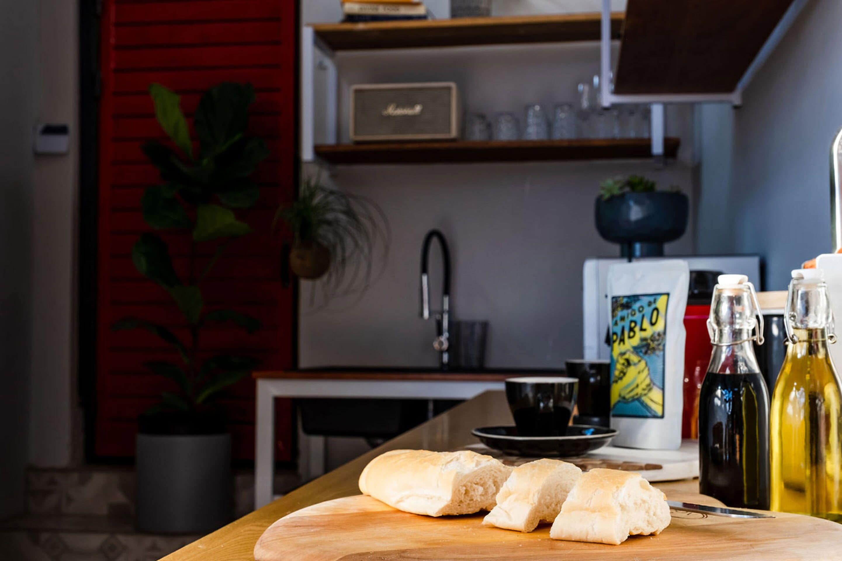 A kitchen with a wooden cutting board, a knife, and a loaf of bread. There are two bottles of olive oil and a book titled "Pablo" on the counter.