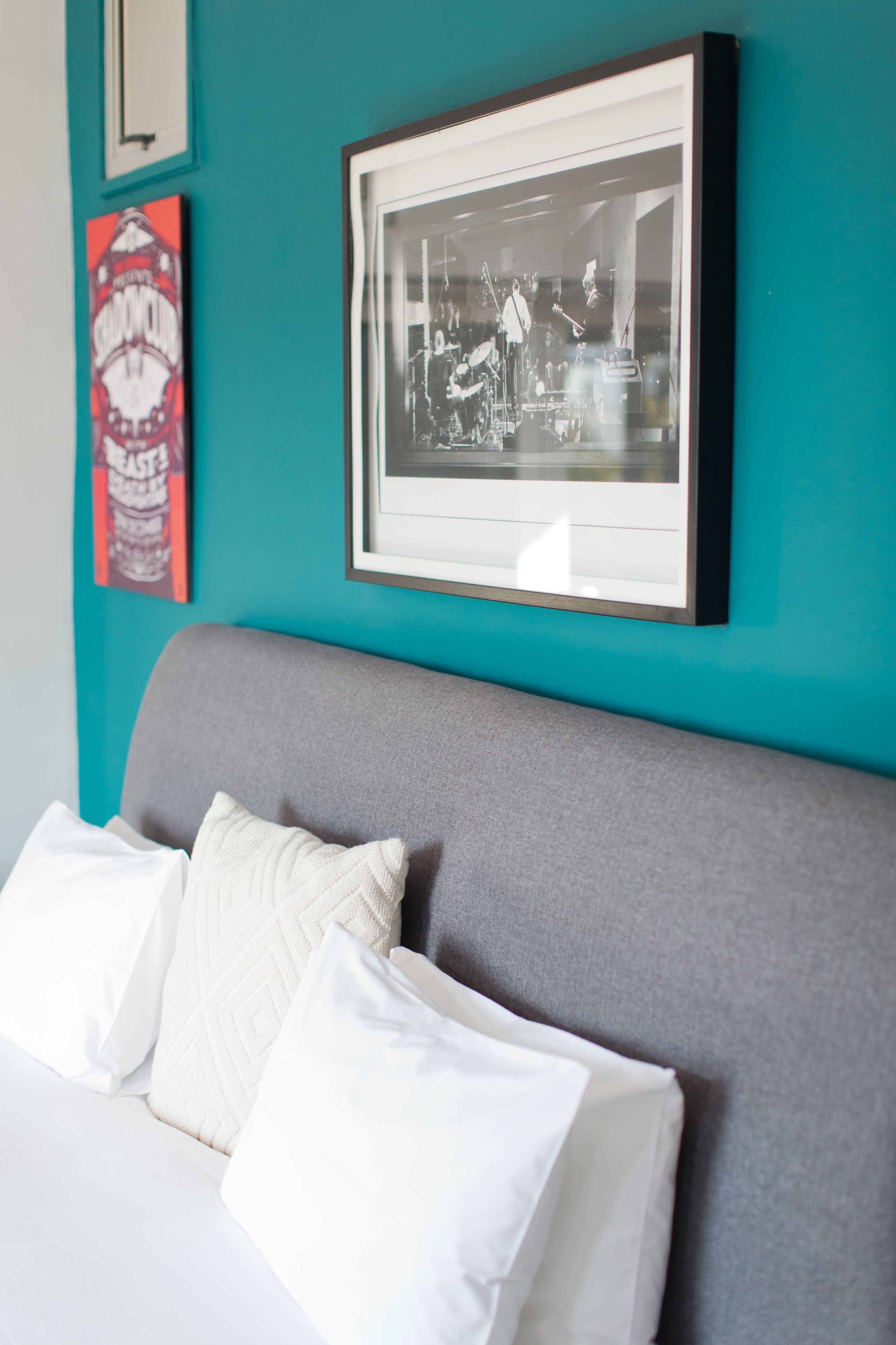 Una cama bien hecha con sábanas y almohadones blancos, una fotografía en blanco y negro enmarcada en la pared, y una pared color verde azulado.