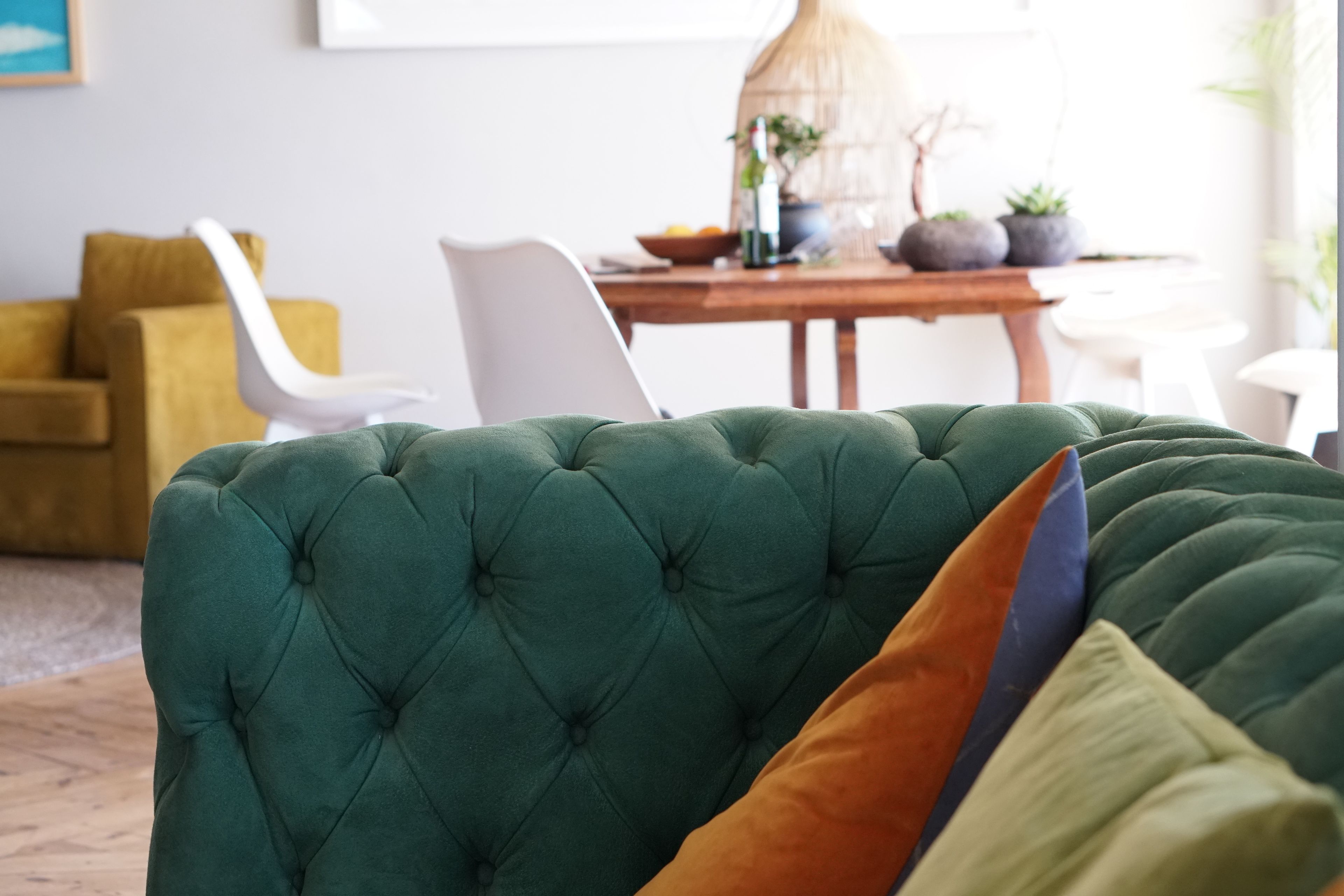 Um sofá verde com tufos e uma almofada, uma mesa de jantar de madeira com cadeiras, e uma planta ao fundo.