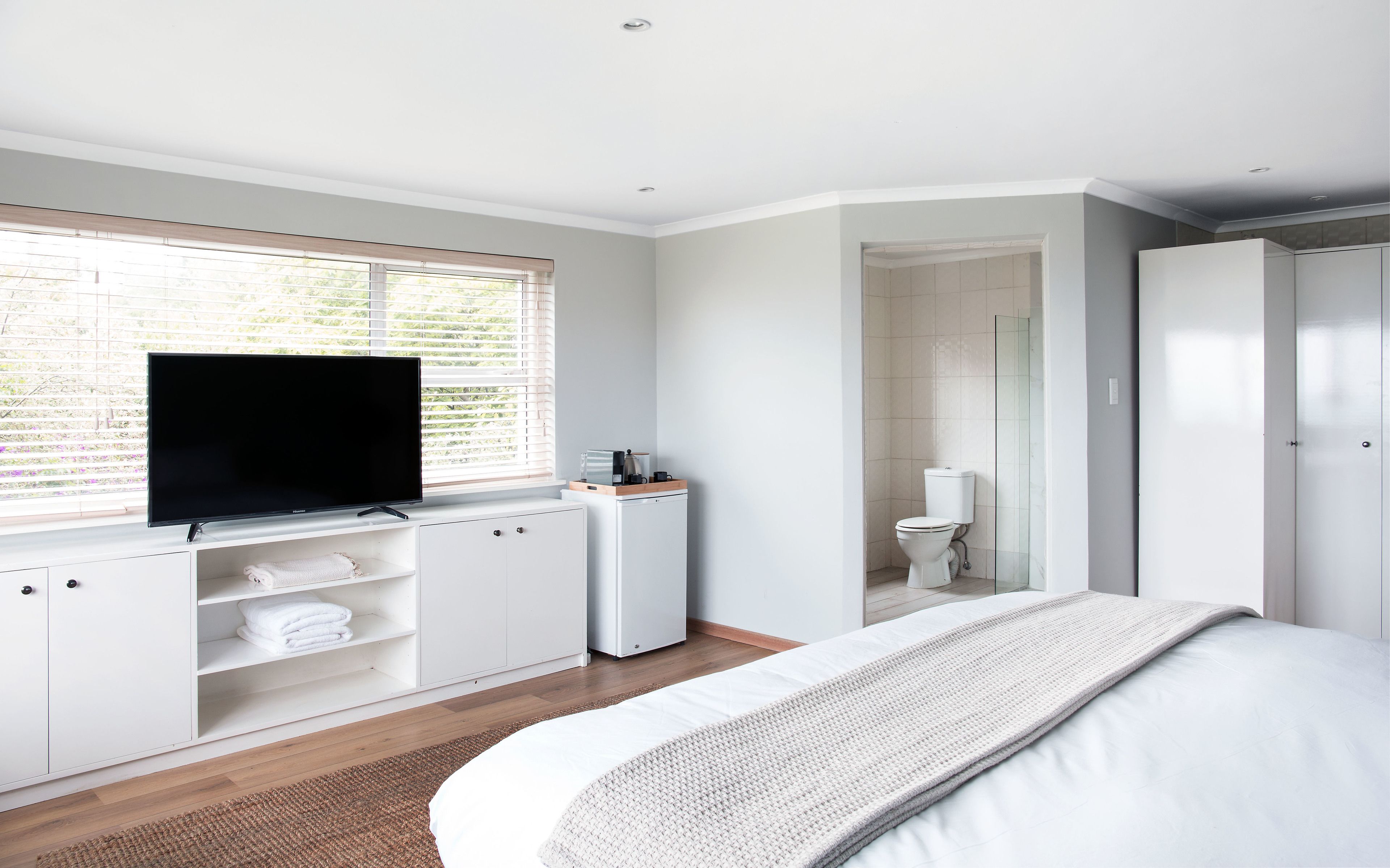 Quarto com cama, televisão sobre um armário branco, banheiro com vaso sanitário e chuveiro, e janela com persianas.
