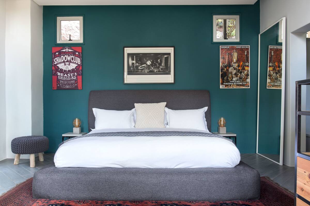 A modern bedroom with a large bed, a grey headboard, and a teal accent wall. There are framed posters and a small stool by the bed.