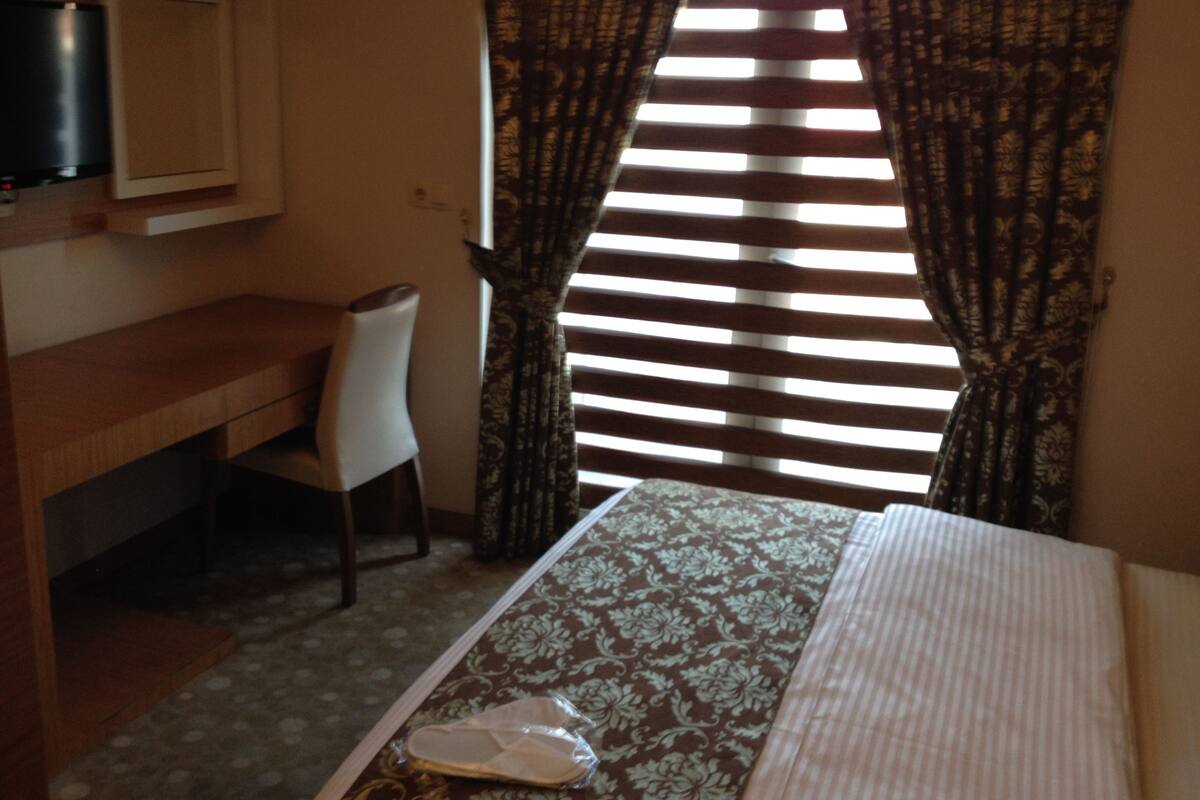 Une chambre d’hôtel avec un lit, un bureau et une chaise. Il y a une fenêtre avec des rideaux et des stores.